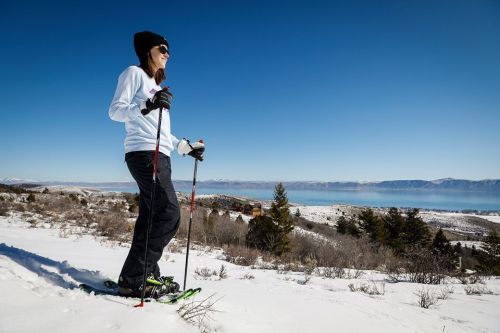 A woman snowshoeing in a snowy landscape, with mountains and a lake visible in the background under a clear blue sky.