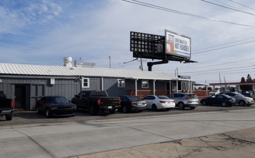 A gray building with a metal roof and several parked cars outside, under a billboard advertising a local business.