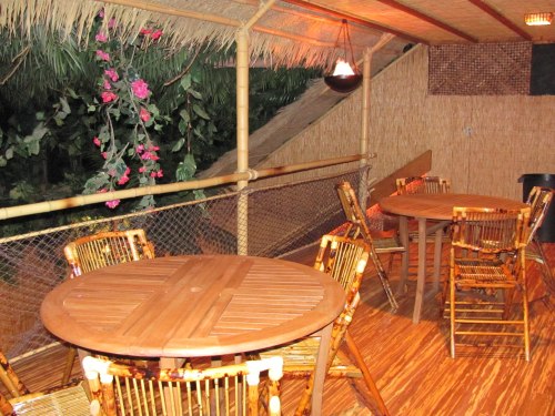 A cozy bamboo terrace with wooden tables and chairs, surrounded by greenery and decorative flowers.