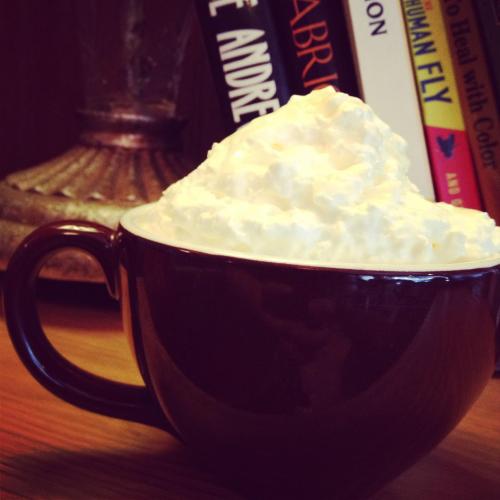 A brown coffee cup filled with whipped cream, placed on a wooden surface with books in the background.