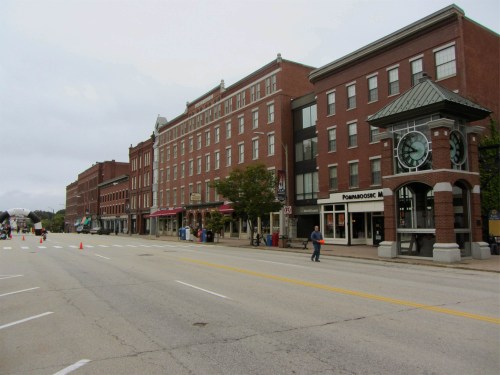 A quiet street lined with brick buildings, featuring shops and a clock tower, under a cloudy sky.