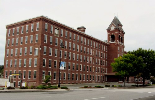 Historic red brick building with a clock tower, surrounded by trees and a parking area, under a cloudy sky.