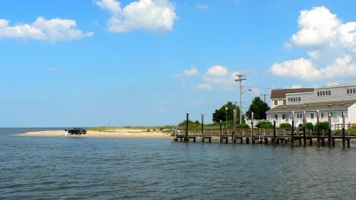 A serene waterfront scene with a sandy beach, a pier, and a house under a blue sky with fluffy clouds.