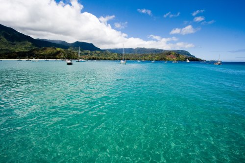A serene turquoise sea with boats anchored, surrounded by lush green mountains and a partly cloudy blue sky.