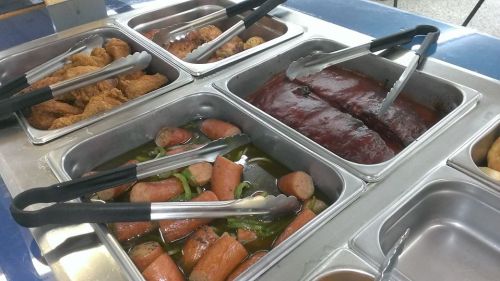 A cafeteria food tray with fried chicken, meatloaf, and sausage with vegetables, all served with tongs.