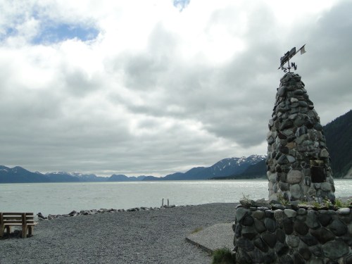 A stone monument with a weather vane overlooks a calm bay surrounded by mountains under a cloudy sky.