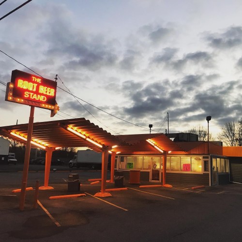 A retro root beer stand illuminated at dusk, with a colorful sign and cloudy sky in the background.