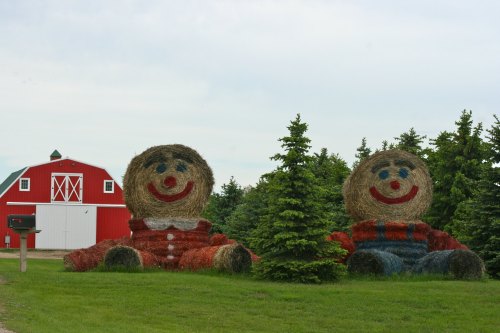 Two large, smiling scarecrow-like figures made of hay bales sit beside a red barn and green trees.