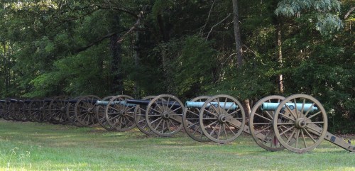 A row of historical cannons on wheels, set against a backdrop of lush green trees.