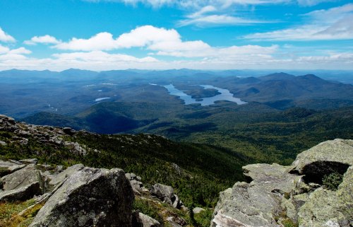 A panoramic view of mountains and lakes under a blue sky, with rocky foreground and lush greenery in the distance.