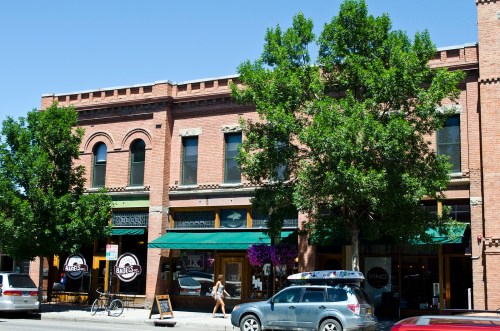 A historic brick building with green awnings, trees, and storefronts, set against a clear blue sky.