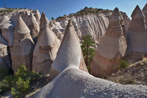 Unique rock formations resembling cones rise from a canyon, surrounded by sparse vegetation and a clear blue sky.