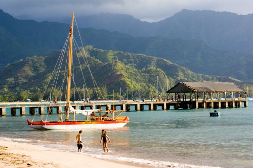 A sailboat anchored near a beach, with two children playing in the water and mountains in the background.