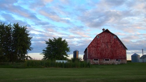 A red barn stands beside trees and a silo under a cloudy sky, with green grass in the foreground.
