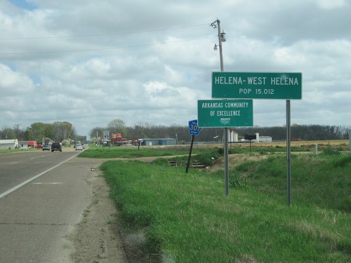 Sign for Helena-West Helena, Arkansas, indicating population and community designation, with a highway in the background.
