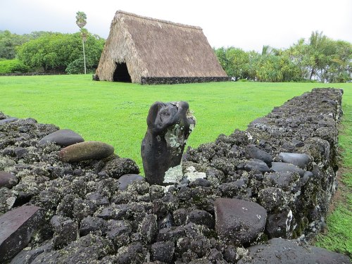 A grassy area featuring a stone wall, a carved figure, and a thatched-roof structure in the background.