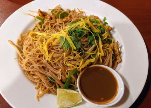 Plate of stir-fried noodles topped with cilantro, fried egg strips, and served with a side of sauce and lime wedge.