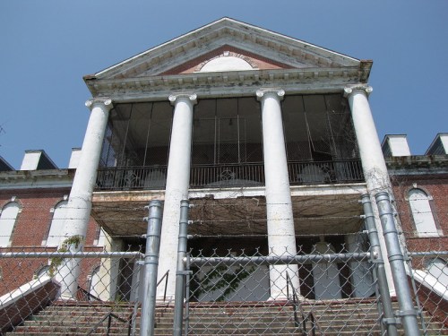 Abandoned building with a grand entrance, featuring tall columns and a fenced-off staircase, under a clear blue sky.