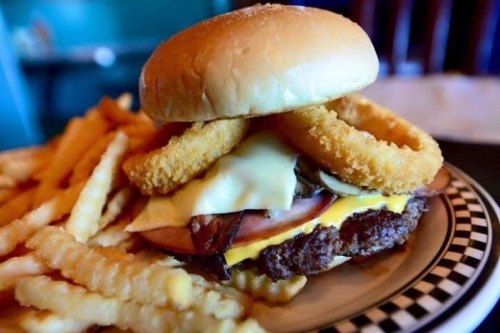 A juicy burger with cheese, ham, and onion rings, served with a side of crinkle-cut fries on a checkered plate.
