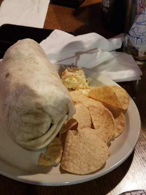 A plate with a large burrito, tortilla chips, and a small serving of coleslaw on a wooden table.