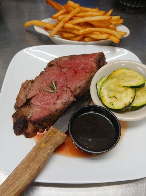 A plate featuring a juicy slice of prime rib with a side of fries and zucchini, accompanied by a small bowl of sauce.