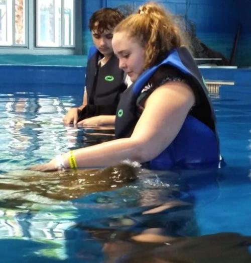 Two people in life vests interact with a large animal in a blue-tinted pool.