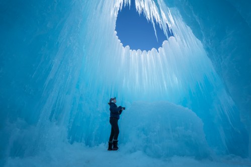 A person stands inside a stunning ice cave, gazing up at icicles hanging from a circular opening above.