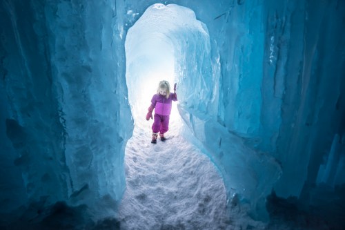 A child in a pink snowsuit stands inside a shimmering ice cave, surrounded by blue ice and snow.