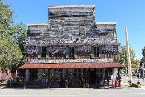 A rustic, weathered country store with a metal roof and wooden facade, surrounded by trees and a clear blue sky.