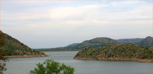 A serene landscape featuring a calm lake surrounded by rocky hills and gentle greenery under a cloudy sky.
