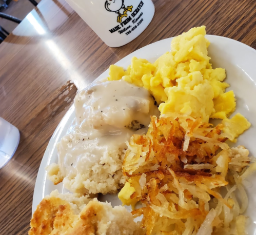 A plate of breakfast featuring scrambled eggs, biscuits with gravy, and crispy hash browns, with a coffee mug in the background.