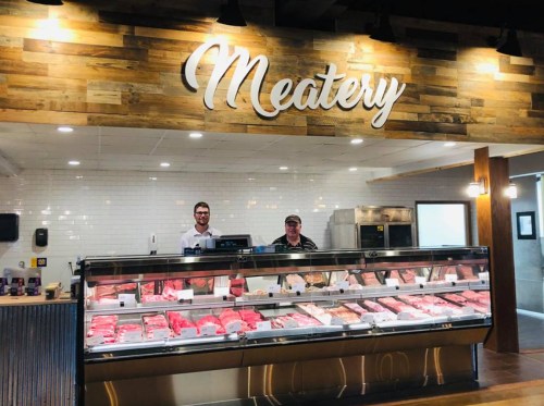 Two butchers stand behind a meat display in a shop labeled "Meatery," showcasing various cuts of meat.