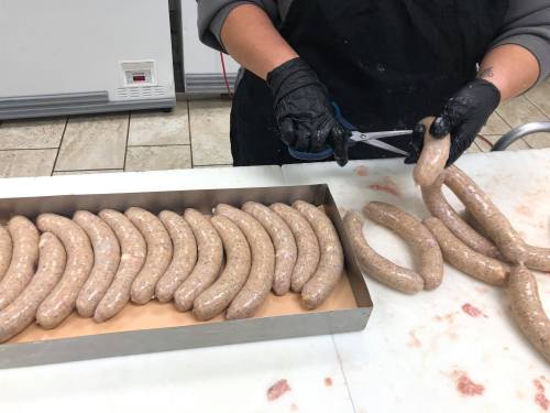 A person in black gloves is cutting sausage links on a countertop, with several uncooked sausages arranged in a tray.
