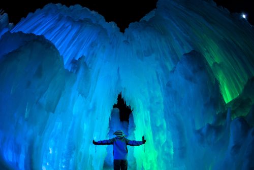 A person stands with arms outstretched in a colorful ice cave illuminated in blue and green lights.