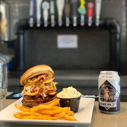 A juicy burger topped with onions and bacon, served with fries and coleslaw, alongside a can of Cosmic IPA.
