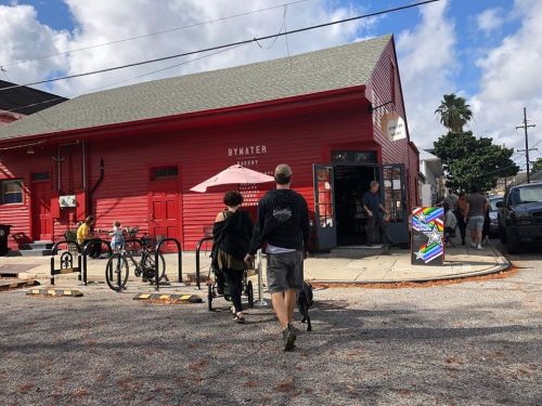 A vibrant red building with people walking outside, bicycles parked, and a colorful sign in front on a sunny day.
