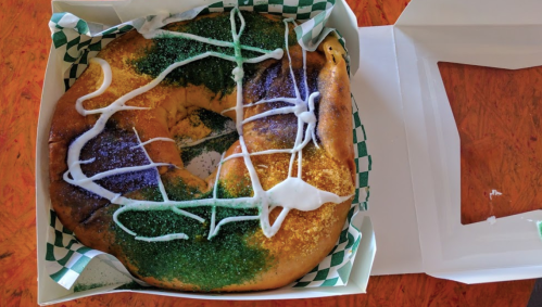 A colorful king cake with green, purple, and gold sprinkles, drizzled with white icing, in a takeout box.