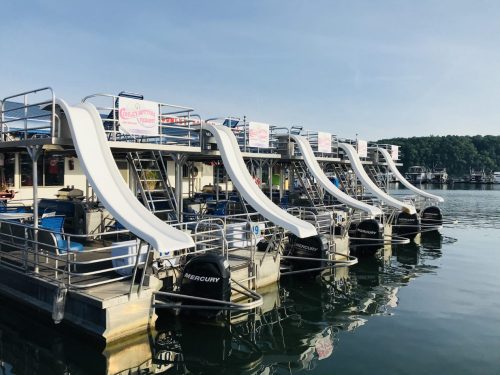 Four houseboats with water slides docked at a calm lake, reflecting the blue sky and surrounding greenery.