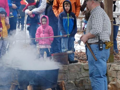 A group of children watches as an adult stirs a pot over a fire, surrounded by smoke and winter scenery.