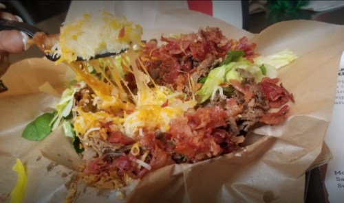 A close-up of a loaded salad with shredded beef, bacon, cheese, and lettuce served in a paper container.