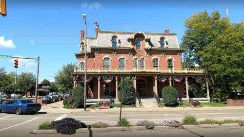 Historic brick building with ornate architecture, surrounded by trees and decorative plants, under a clear blue sky.