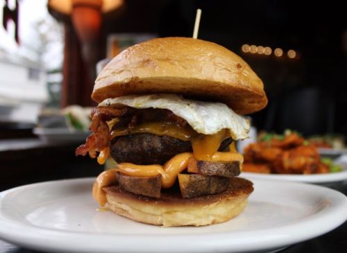 A stacked burger with bacon, cheese, a fried egg, and potato wedges on a toasted bun, served on a white plate.