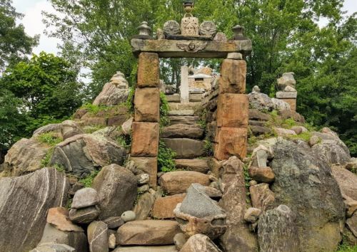 Stone archway leading up a rocky path, surrounded by greenery and adorned with sculptures.