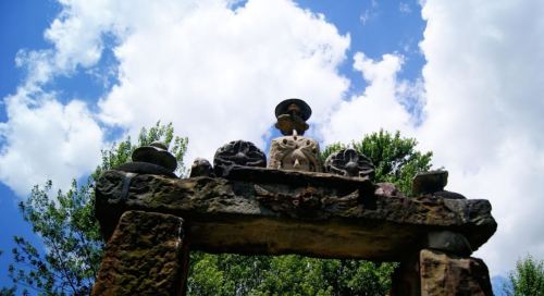 Ancient stone archway with carvings, surrounded by trees and a blue sky with fluffy clouds.