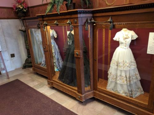 Three elegant dresses displayed in glass cases, showcasing intricate designs and vibrant colors in a museum setting.