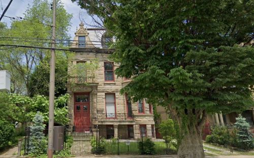 Victorian-style house with red doors, surrounded by greenery and a tall tree, set behind a wrought iron fence.