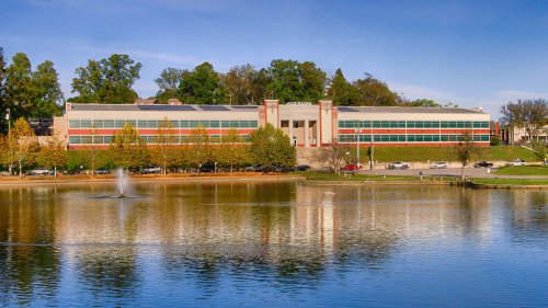 A modern building with red and green accents reflects in a calm lake, surrounded by trees and a clear blue sky.