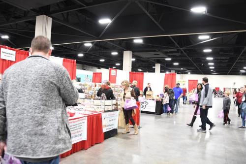 A busy indoor market with vendors at tables, red curtains, and shoppers browsing products.
