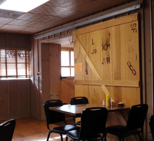 A wooden interior with a table and chairs, featuring a sliding door marked with letters and a bottle of mustard on the table.