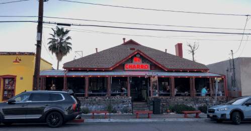 A restaurant named "El Charro" with a rustic exterior, outdoor seating, and palm trees, set against a twilight sky.
